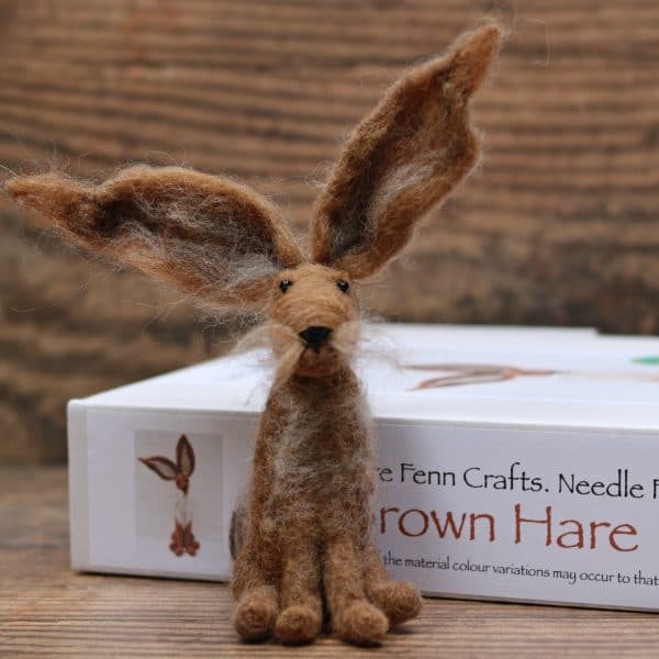 Image shows a cute needle felted brown woodland hare. It is sitting in front of a white box that says, Hare Needle Felting Kit. The background is a warm wood effect.