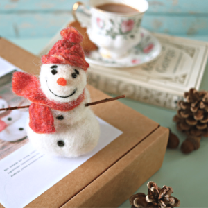 Image shows a cute needle felted snowan with bright red and orange har, sticks for arms, sat on a needle felting kit.