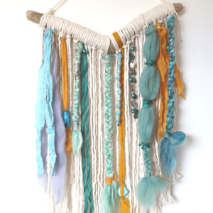 Image shows a boho style wall hanging hung from a piece of driftwood. Colours are calming blues, aqua and orange. Fabrics are macrame rope, wool tops, beads, and ribbons