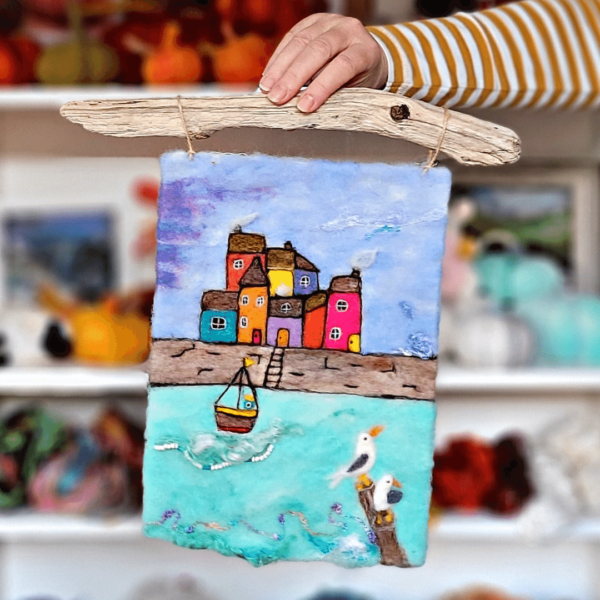 Image shows a needle felted seaside scene with harbour, brightly coloured houses, boat and two seagulls. Text on image reads: New picture kit. cornish seascape. Learn to paint with wool.